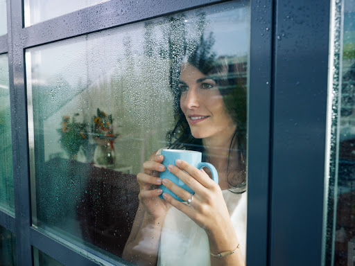A woman holding a cup of coffee as she looks outside a window with condensation on it.
